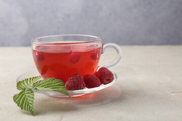 Tasty raspberry tea in glass cup, berries and green leaves on light grey table, closeup. Space for text