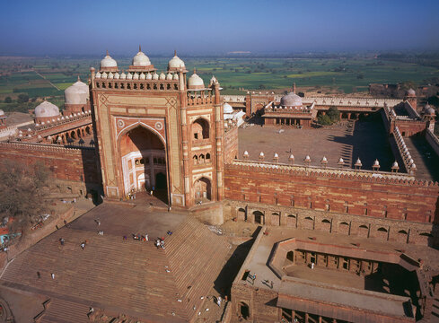Aerial view of red sandstone walls of the Jama Masjid mosque, with domes and arched gateways, against a backdrop of green fields, Fatehpur Sikri, India.