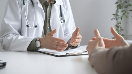 Doctor wearing lab coat and stethoscope is gesturing with hands while explaining diagnosis to...
