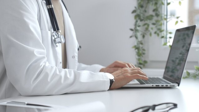 Male doctor wearing a white coat and stethoscope is typing on a laptop in a bright medical office, showcasing the intersection of healthcare and technology, close up. Medicine and health care concept