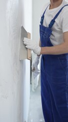 Man construction worker wearing protective gloves and blue construction coveralls, spreading plaster smoothly across wall using professional drywall taping knife