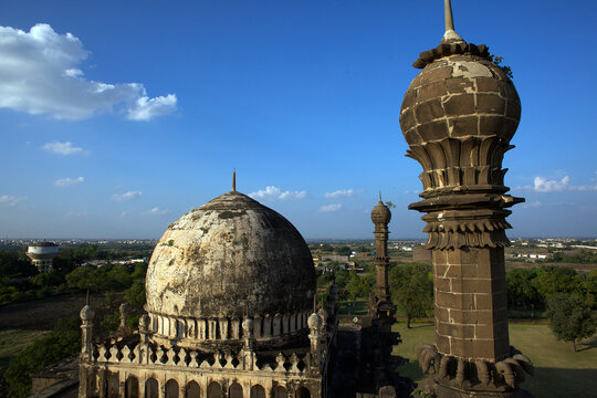 Aerial view of the weathered dome and minaret of Ibrahim Roza against a backdrop of a vibrant blue sky and distant landscape, Vijayapura, Karnataka, India.