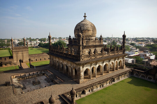 Aerial view of the majestic Ibrahim Rouza complex with intricate arches and a bulbous dome, framed by green lawns and a distant cityscape, Vijayapura, Karnataka, India.