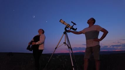 Amateur astronomers using telescope and binoculars for watching stars, Moon, planets and other celestial objects from a countryside field in nature.