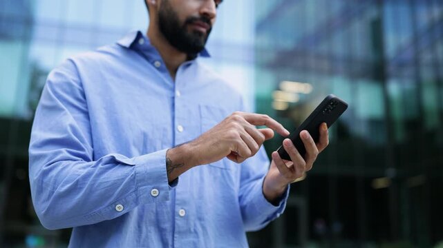 A smiling man with a beard uses a smartphone, showcasing modern technology in an urban setting with a blurred background.