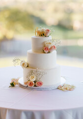 Beautiful three-tiered white wedding cake outdoors. Creamy delicate holiday cake decorated with pink roses and the word love on top. Wedding cake stands alone isolated on celebration