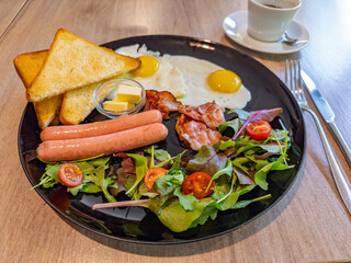 Classic full English breakfast with fried eggs, sausages, bacon, toast, and fresh salad on a black plate.Delicious morning meal in a restaurant