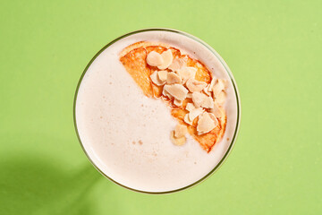 A bright orange cocktail with a foam top is garnished with a dried orange slice and almond flakes, served in a coupe glass on a green background.