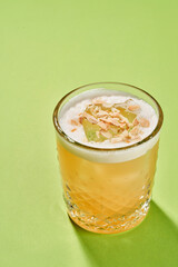 A yellow cocktail with a foam top is garnished with a translucent fruit piece and almond flakes, served in a highball glass on a green background.