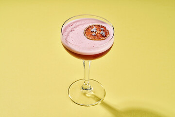 A stylish pink cocktail with a foam top is garnished with a dried orange slice and delicate flowers, served in a coupe glass on a yellow background.