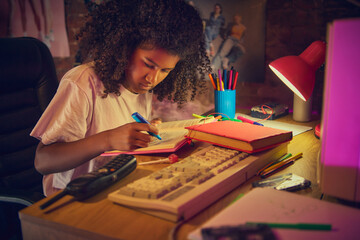 Fototapeta premium Teen girl carefully highlights text in open book at wooden desk with vintage computer. Concept of study process, organization, and productivity in a nostalgic workspace.