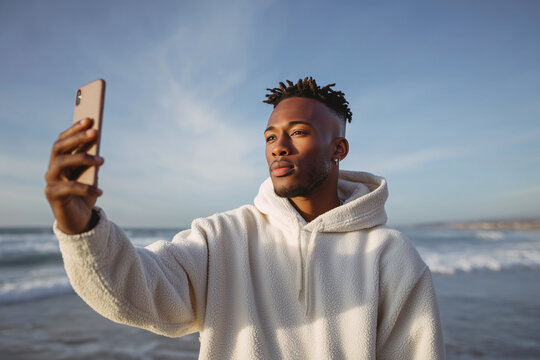 Young man in white hoodie taking a selfie by the ocean during golden hour with calm waves and clear blue sky