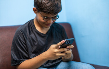 Fototapeta premium Asian guy teenager sitting on sofa at home relax using mobile phone app typing text browsing message, gen z checking social media on webpage, happiness lifestyle digital communication concepts