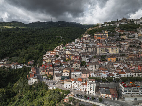 Aerial view of a medieval town cascading down a hillside, with terracotta roofs contrasting against the lush green forests under a brooding sky, Morcone, Benevento, Italy.