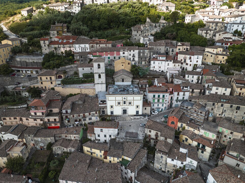 Aerial view of sun-kissed rooftops and the towering spire of the church create a tapestry of terracotta and cream, nestled amidst verdant hills, Morcone, Benevento, Italy.