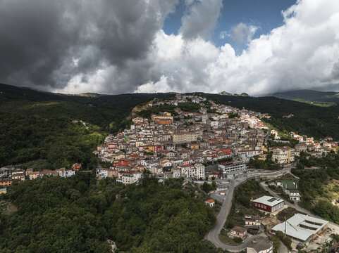 Aerial view of sunlit buildings nestled amidst verdant hills under a dramatic sky, creating a picturesque contrast of light and shadow, Morcone, Benevento, Italy.