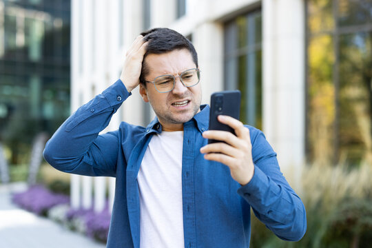 A man looks frustrated while using his smartphone outdoors. The building backdrop and his expression suggest a displeasing or confusing interaction with technology or communication.