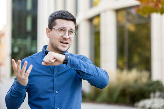 A man wearing glasses and a blue shirt interacts with a smartwatch, gesturing animatedly while standing outdoors near a modern office building.