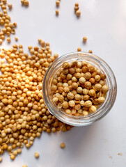 dried coriander seeds spill from a glass jar onto a white surface, creating a beautiful pattern with warm, earthy tones.