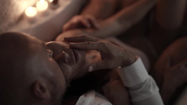 The man gently touches the woman's face, she is relaxed in his arms, candles flicker behind on the side of the tub.