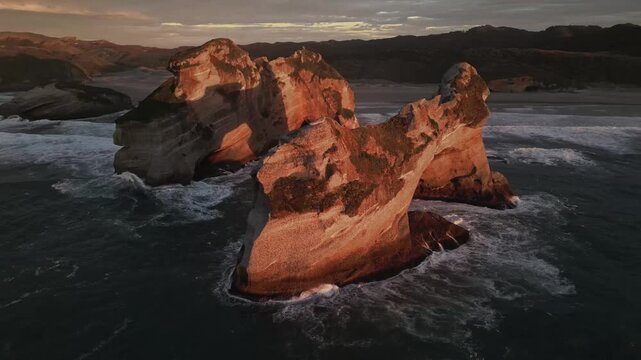 Aerial view of Wharariki Arch rocks sculpted by the ocean, the sun paints the rock formations in warm shades of orange against the dark blue sea, Puponga, New Zealand.