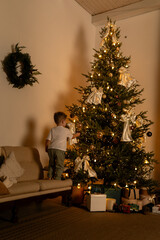 Child decorating Christmas tree with ornaments and lights in cozy living room
