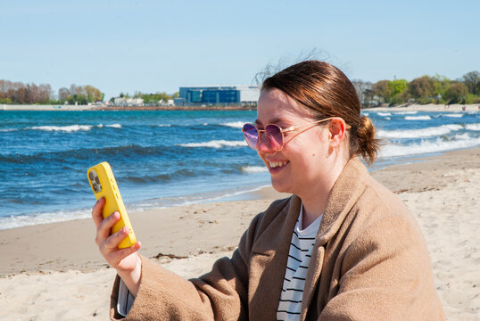 Smiling woman in beige coat and pink sunglasses holding smartphone during video call on sandy beach near ocean waves under sunny blue sky representing travel, communication and digital nomad lifestyle