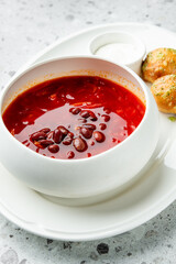 Red bean soup with vegetables served in a bowl, accompanied by garlic bread rolls and sour cream on a white plate over a speckled gray surface