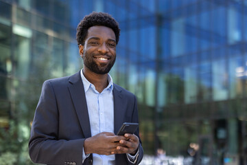 Confident businessman standing outdoors in front of a modern glass building using a smartphone. Dressed in formal wear, he exudes professionalism while engaging with his mobile device.