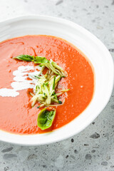 Creamy tomato soup in a white bowl, garnished with shredded zucchini, basil leaves, and cream drizzle, served on a speckled gray countertop, top view