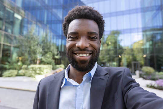 Confident businessman smiles as he takes a selfie, standing outside a contemporary glass office building.