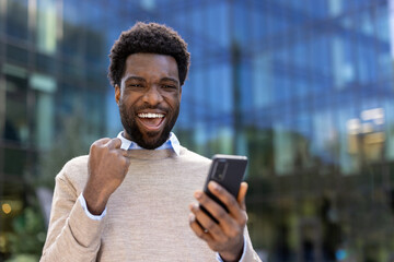A cheerful man dressed casually is expressing excitement while using a smartphone outside a modern glass-covered building. The photo captures joy, technology, and a professional office environment