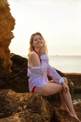 Cute blonde in red swimsuit and white shirt sits on rock against sea background.