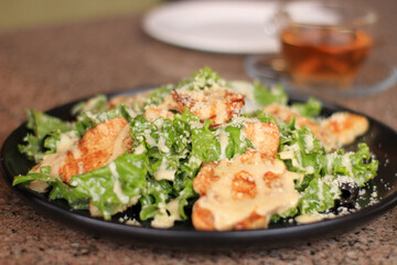 A black plate holds a fresh Caesar salad topped with grilled chicken slices, drizzled with creamy dressing, and sprinkled with grated cheese, set against a blurred background.