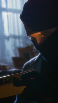 Vertical shot of man wearing black balaclava holding shiny golden bar while admiring it during housebreaking