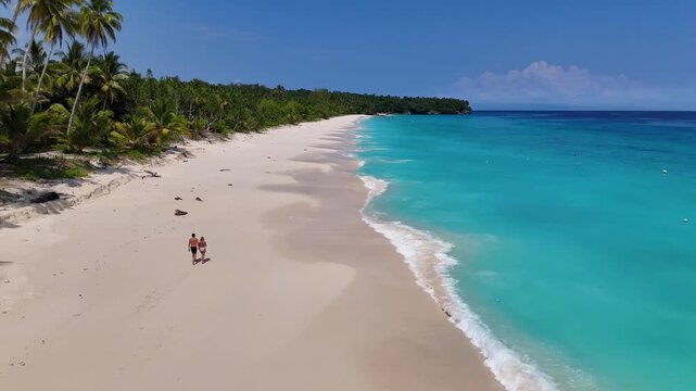 4K drone video flying over a couple walking along a tropical white sand beach in Indonesia. This is Mandel Beach in Sulawesi, a stunning white sand beach lines with coconut trees and a blue ocean.