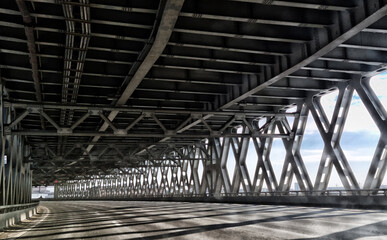 View from inside a metal truss bridge in St. Petersburg. Sunlight filters through the structure, creating a dramatic pattern of shadows on the asphalt.  
