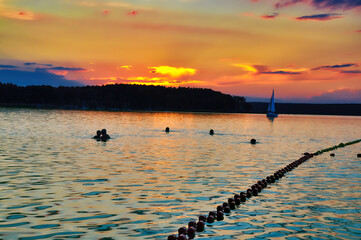 Niesulice - Polska Piękny zachód słońca odbijający się w spokojnej wodzie, pływacy cieszący się krajobrazem i żaglówka w oddali tworzą atmosferę spokoju i ukojenia, symbolizującą lato i wypoczynek. © Pol Badger