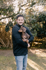 Man with black hair and wearing black holding and cuddling a brown dachshund. Love your pet, dog, companion