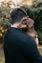 Man with black hair and wearing black holding and cuddling a brown dachshund. Love your pet, dog, companion