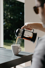 Man pouring filter coffee from a french press into a white cup at an outdoor table
