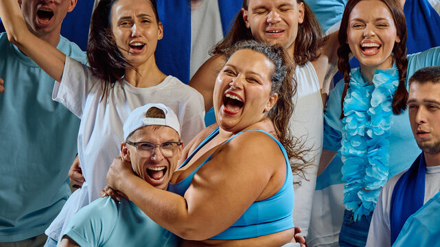 Plus size woman in blue sports bra and shorts hugs male friend in cap while laughing among cheering crowd in blue and white colors. Concept of emotional unity, friendship, and celebration.