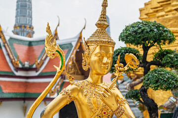 Golden Kinnari statue with ornate headdress and jewelry at the Grand Palace complex in Bangkok, Thailand, cultural tourism.