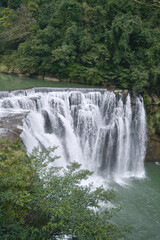 Fototapeta premium Majestic waterfall cascading down natural rock formations into a serene river, surrounded by lush green forest, natural beauty.