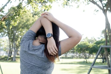 Adult woman stretching arm and shoulder in a park wearing a smartwatch and earphones, focusing on fitness and wellness.