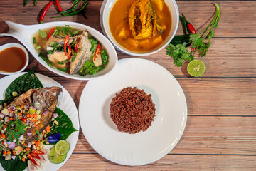 Traditional Thai fish dishes on a wooden table, featuring fried whole fish, fish stew, fish curry, and brown rice with fresh herbs. Authentic Asian cuisine.