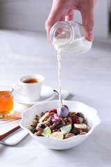 Hand pouring milk into a white bowl filled with healthy breakfast cereal mixed with fresh fruits and nuts on a bright table. Healthy Breakfast.
