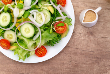 Fresh green salad with cucumber, tomato, red onion, and avocado on a white plate next to salad dressing on a wooden table. Healthy eating concept.