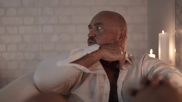 The man looks warily to the side, leaning slightly on his arm, surrounded by candles.