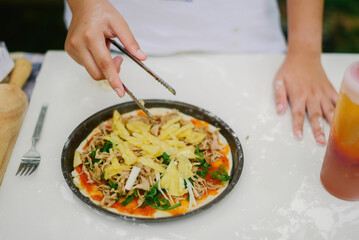 Kid's hands adding pineapple and meat toppings to a homemade pizza with tomato sauce and greens on a white kitchen counter, cooking concept.